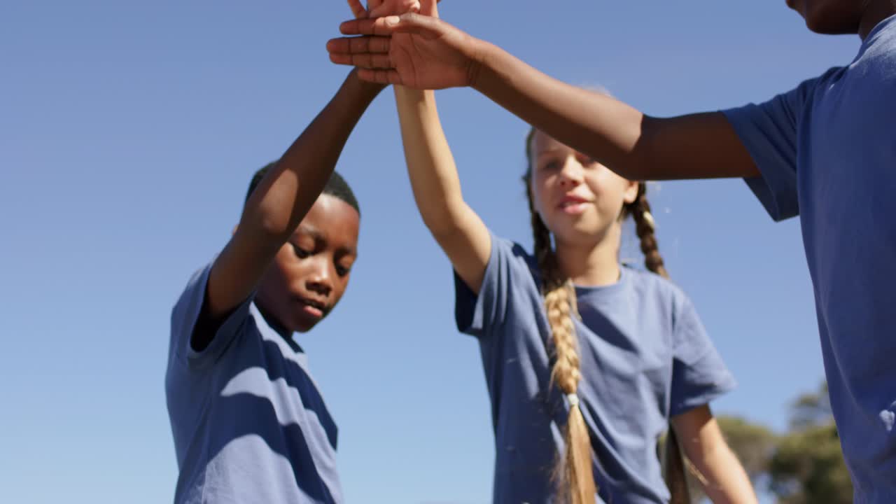 Diverse children in blue tees after girl's braid cue, stacking hands, lifting arms cheering at park