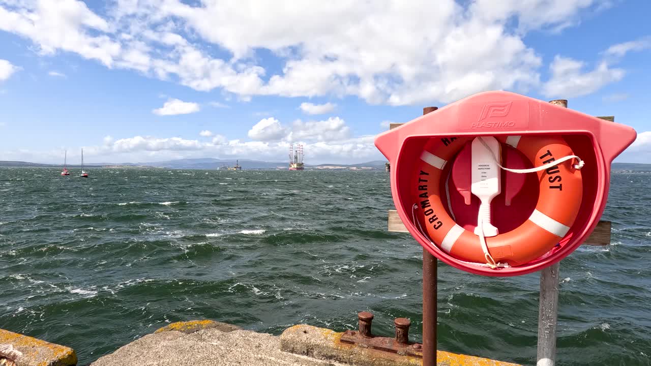 Alternating shots reveal a red lifebuoy station and rough sea under a partly cloudy sky, with smooth lateral camera movement and natural daylight