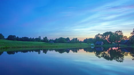 Farbenfroher-Zeitraffer-Von-Der-Morgendämmerung-Bis-Zum-Sonnenaufgang-Eines-Bauernhauses-Am-Seeufer,-Wobei-Sich-Der-Himmel-Im-Wasser-Spiegelt