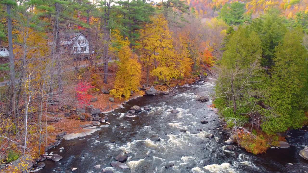 Mont Tremblant, Québec, Canada, with a peaceful river flowing through a vibrant autumn landscape, colorful foliage lining the banks, and cozy houses nestled near the water's edge. Drone view