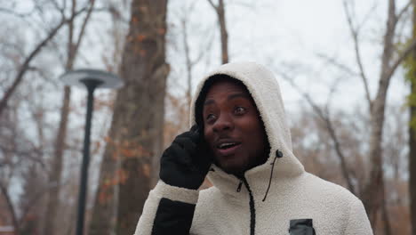 young man conversing over phone, smiling and engaged in lively conversation, wearing a white hoodie and black gloves, with blurred background of trees, street light poles in urban setting