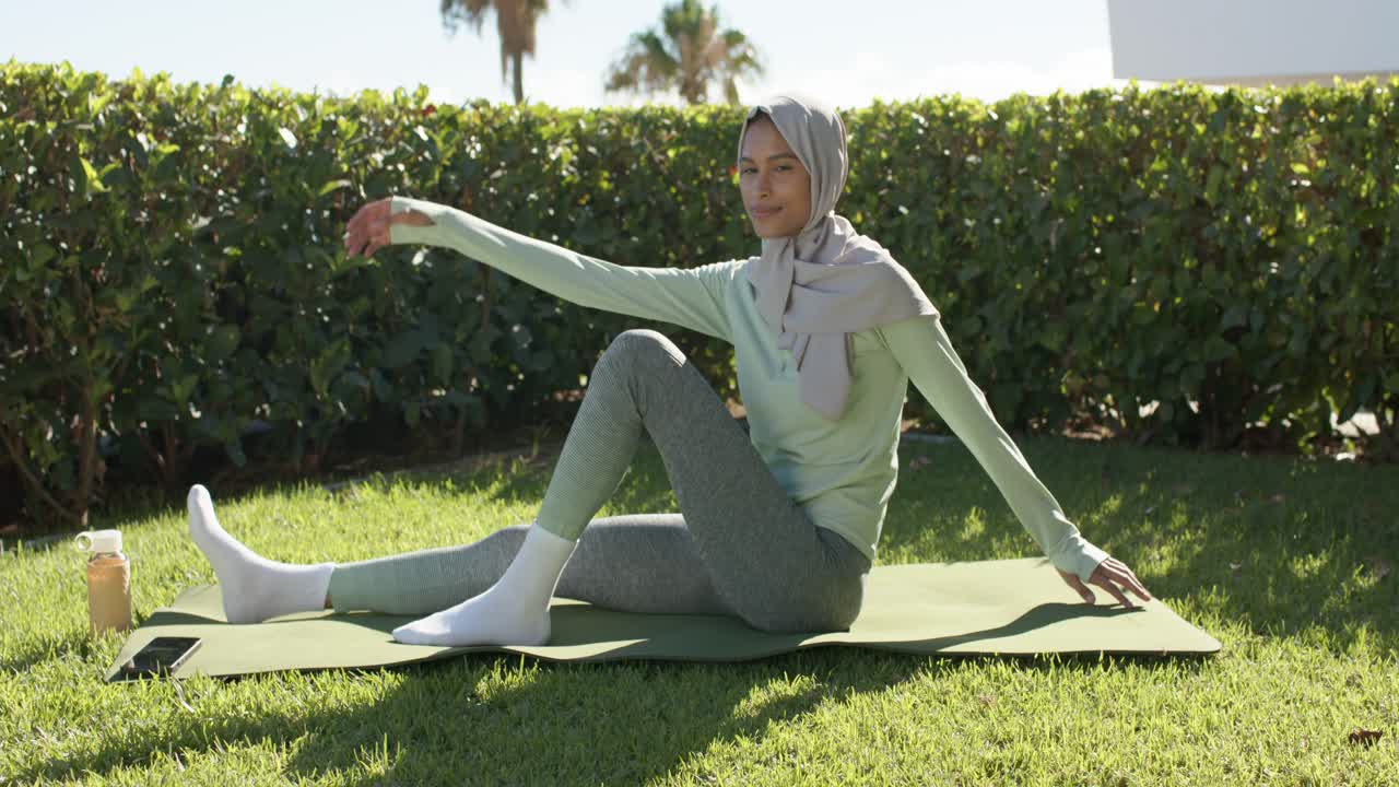 African American woman in hijab legs extended doing twist on green mat on lawn, bottle, stretch