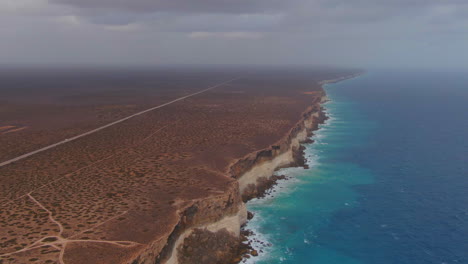 High-panning-drone-shot-of-a-camper-van-driving-over-an-empty-road-next-to-the-high-cliffs-in-a-barren-environment