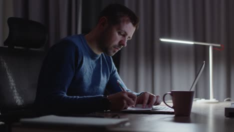 tired male entrepreneur working with writing ideas on a note pad， checking his wrist watch, rubbing sleepy face in dark home office workplace with laptop