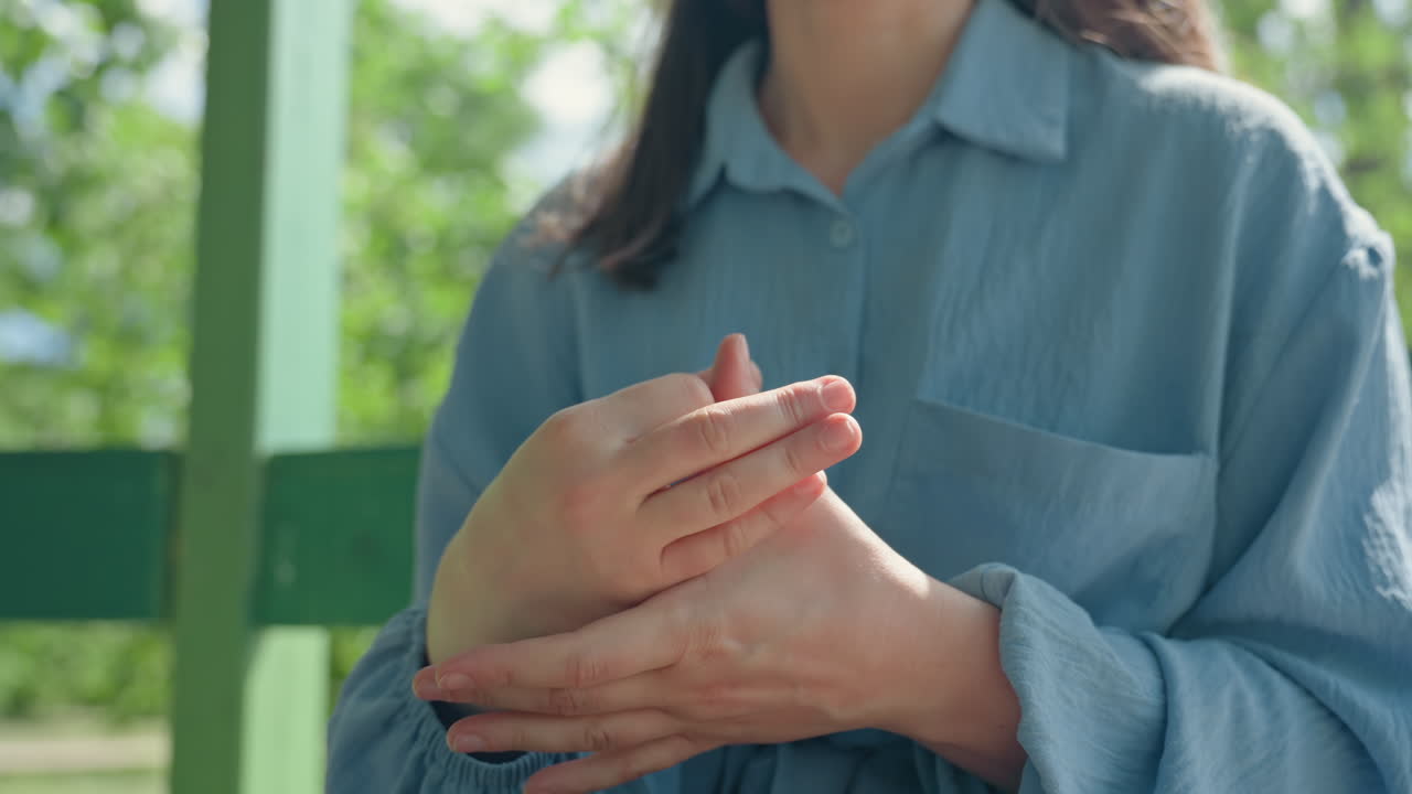 백인 여성이 무릎 위에서 손과 손가락을 비비고, 파란색 셔츠 소매가 약간 당겨진 채 실외 공원 벤치에 앉아 있는 장면, 긴장된 기대감과 초조한 에너지, 햇빛과 부드러운 그림자, 보이는 대처 제스처