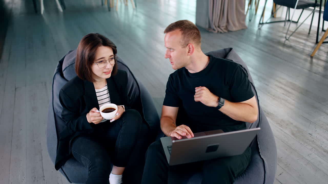 Co-working people sit in soft bean bag chairs. Man holds a laptop commenting something to the girl. Lady is drinking coffee. High angle view.