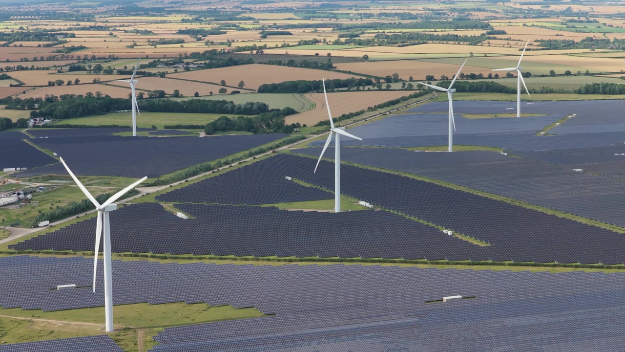 Aerial view of large wind turbines and solar panels covering rural farmland to produce clean energy during a bright afternoon day in England with green hills in the distance