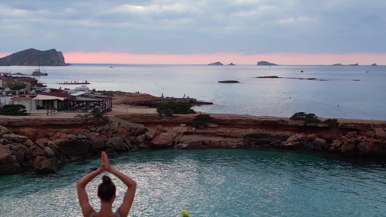 Majesteettinen ilmakuva dronella: Jooga-asennossa (puu) oleva tyttö auringonlaskussa Ibizan saaren kalliorannalla Espanjassa, ylilentokuva.