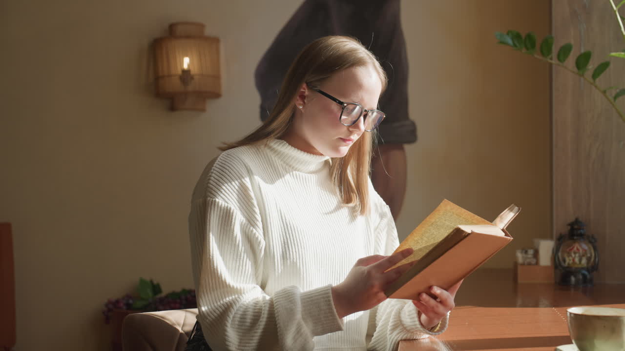Lady in white sweater reading book near window as natural sunlight illuminates pages, creating warm inviting atmosphere with potted plant and cup on wooden table