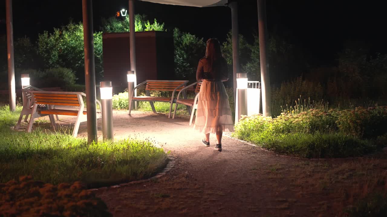 joven hermosa con cabello largo camina en el jardín a la glorieta iluminada con bancos y lámparas por la noche