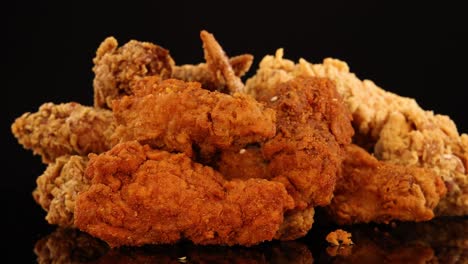 a rotating display of assorted crispy fried chicken wings, drumsticks, and nuggets on a reflective black surface, illuminated by warm studio lighting
