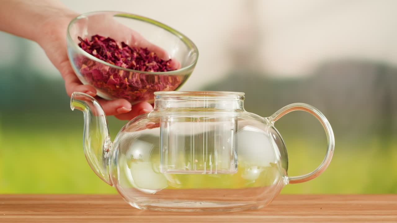 Person adding dried rose petals into a glass teapot for brewing tea