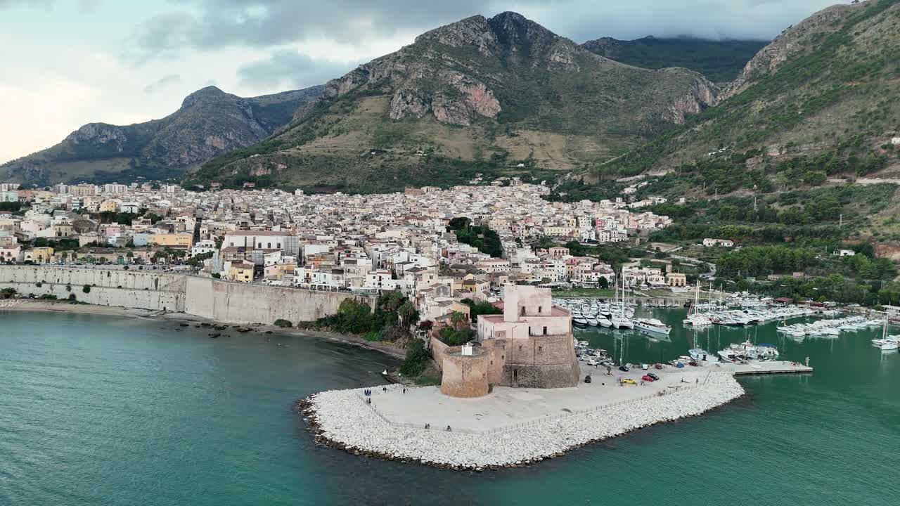 Aerial fly-back and left reveal over Castellammare del Golfo in Sicily, showing the waterfront, castle, and mountain backdrop in soft coastal light