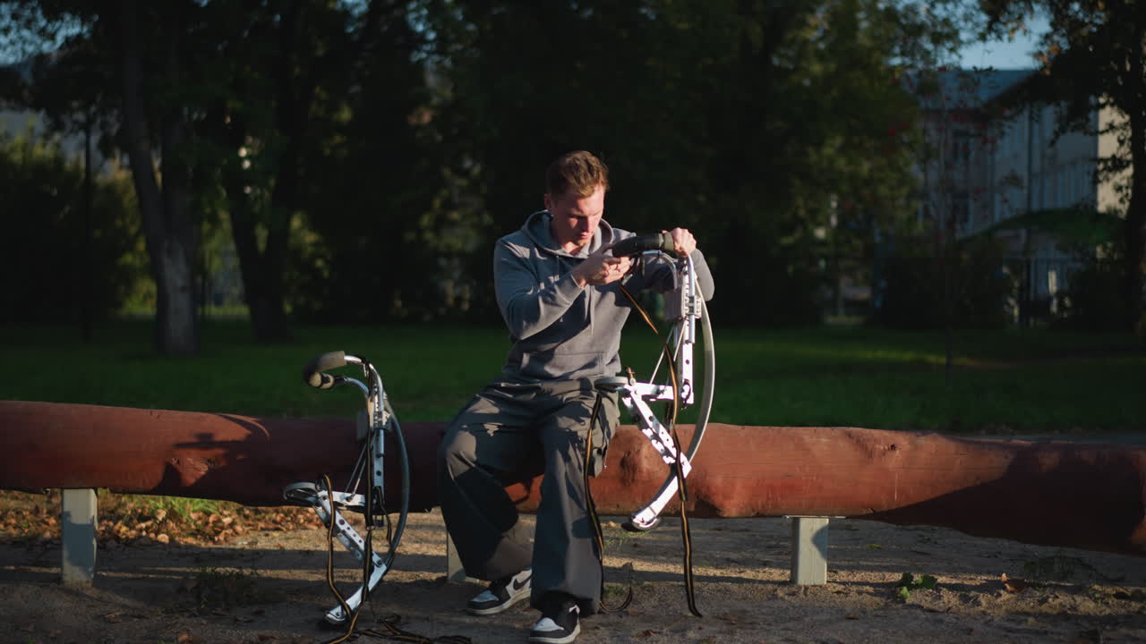 Person in gray hoodie and dark pants focused on adjusting stilts in sunny outdoor setting surrounded by lush greenery. Concentrated expression shows attention to detail, with natural light