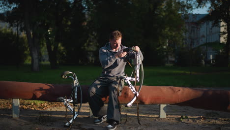 person in gray hoodie and dark pants focused on adjusting stilts in sunny outdoor setting surrounded by lush greenery. concentrated expression shows attention to detail, with natural light