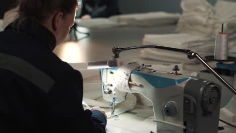 la vista desde atrás muestra a una mujer con ropa de trabajo cosiendo tela blanca en una máquina de coser en una fábrica textil