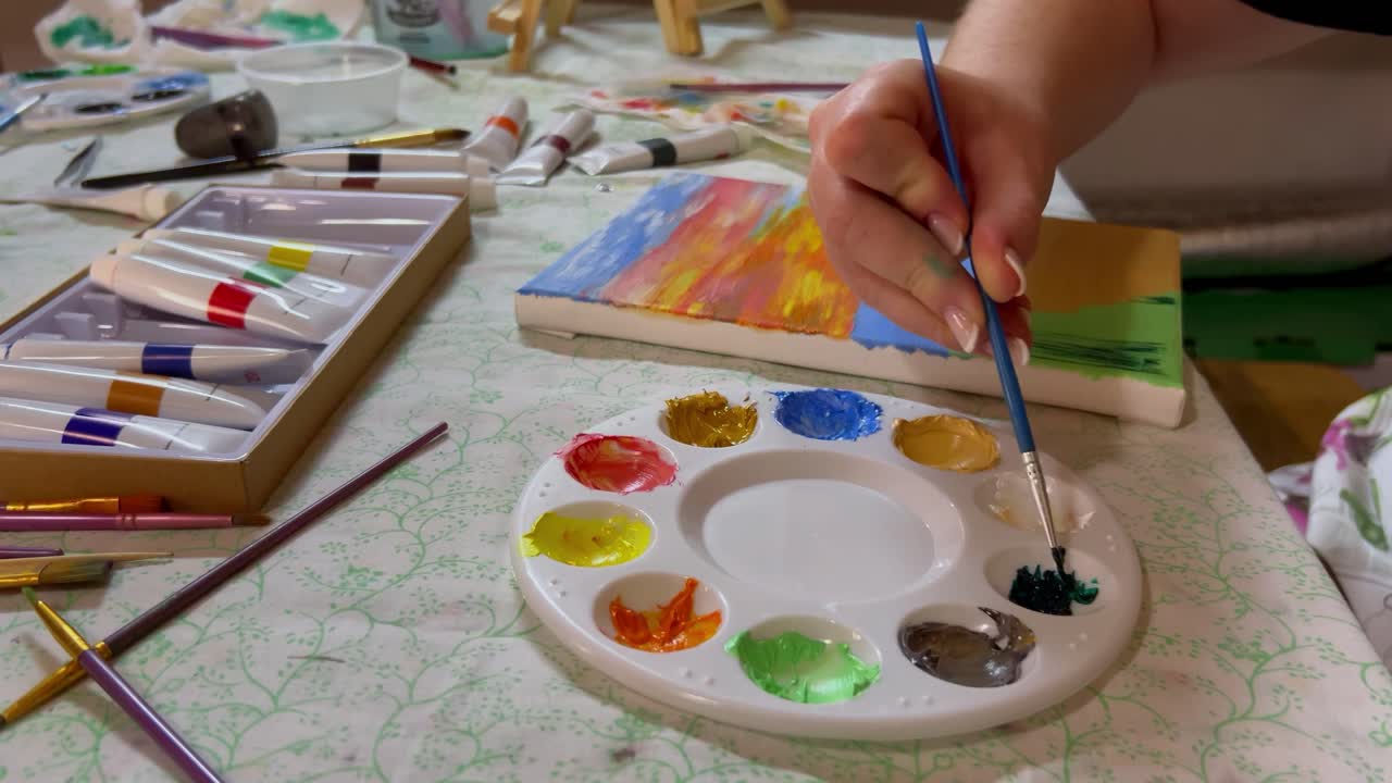 A woman's hands are visible as she uses dark green oil paint to paint the plants on her beach sunset oil painting. She uses a thin brush and long brush strokes