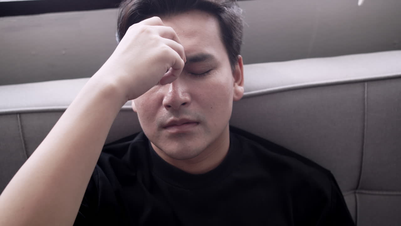 Young man slouched on the sofa, feeling stressed and overwhelmed with a headache due to work exhaustion, societal pressure, and the impact of insufficient rest on mental health.