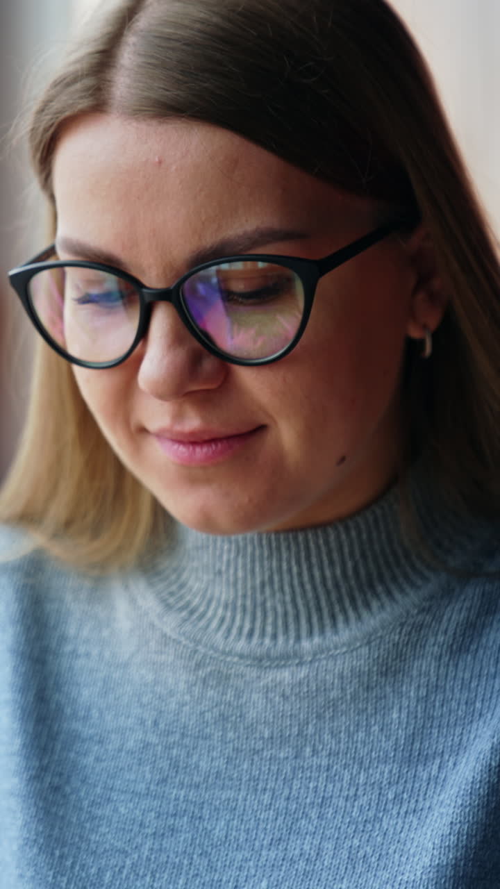 Calm positive Caucasian woman wearing glasses sits at desk. Lady stops typing on her laptop, takes off glasses and smiles to camera. Vertical video