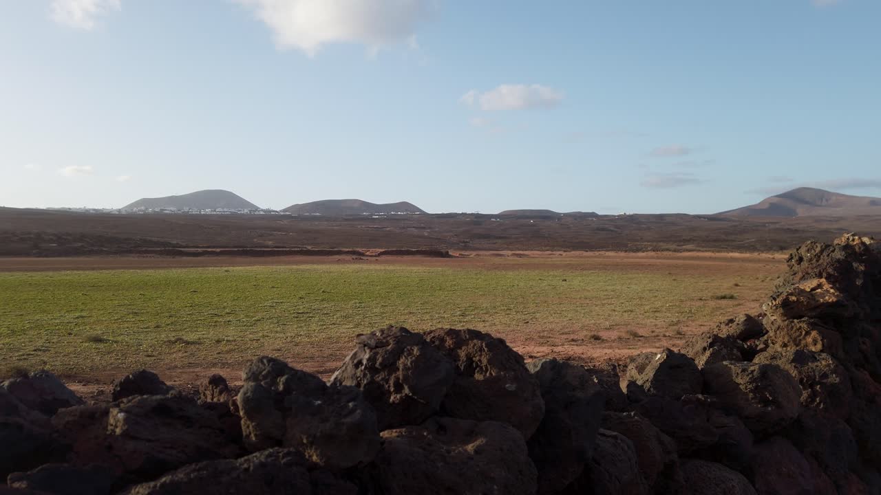 Verdant plain bordered by rugged volcanic rocks with majestic mountain silhouettes under expansive sky in lanzarote's serene volcanic landscape, canary islands, spain