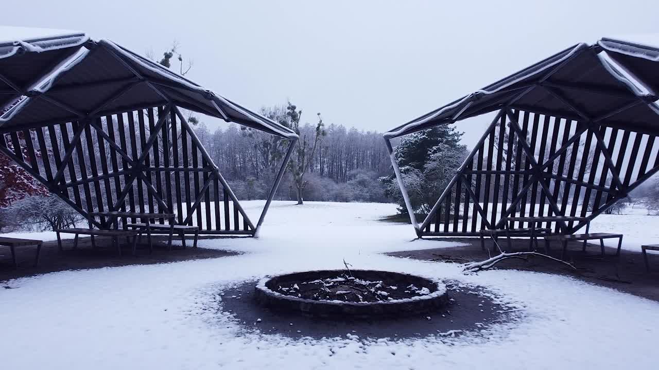 punto de vista a través de un lugar de picnic con cobertizos modernos, desierto en invierno y luego volando para mirar desde arriba los alrededores: prado cubierto de nieve, árboles, bosque y lago