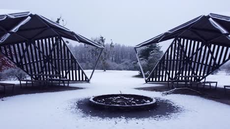 Pov-through-picnic-spot-with-modern-sheds,-deserted-in-the-winter-and-then-flying-to-look-from-above-at-surroundings:-snow-covered-meadow,-trees,-forest,-and-lake