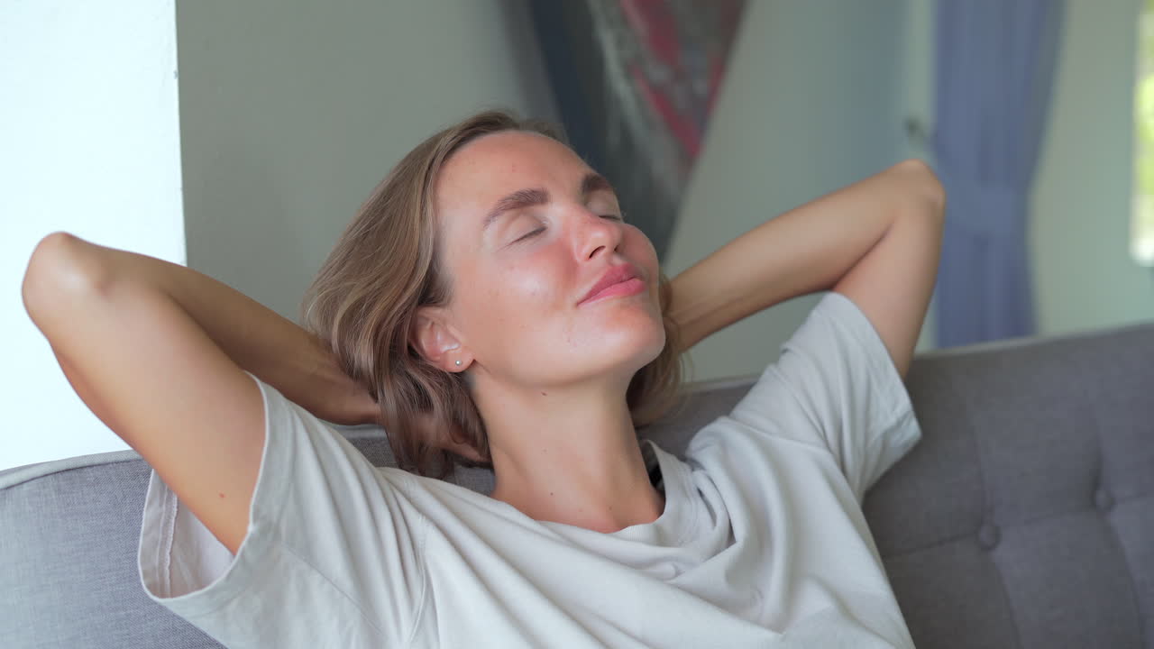 Calm young woman dozing daytime nap on couch with closed eyes. Girl closed hands behind hand breathing fresh air resting in comfortable sofa at home
