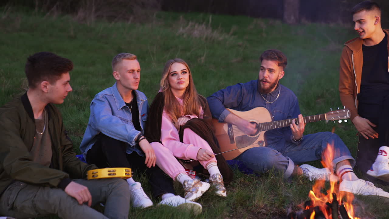 Merry singing near the campfire on beautiful summer evening. Young people sit on the grass, fry marshmallows and sing songs.