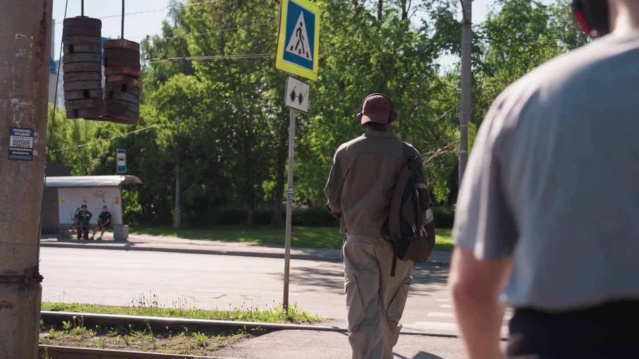 Man wearing cap and carrying backpack walks steadily along sunlit sidewalk near train tracks passing by another man with headphones. scene includes green trees pedestrian signs on poles