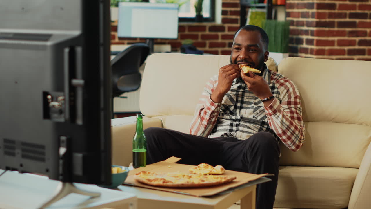 Young male adult eating slice of pizza at tv