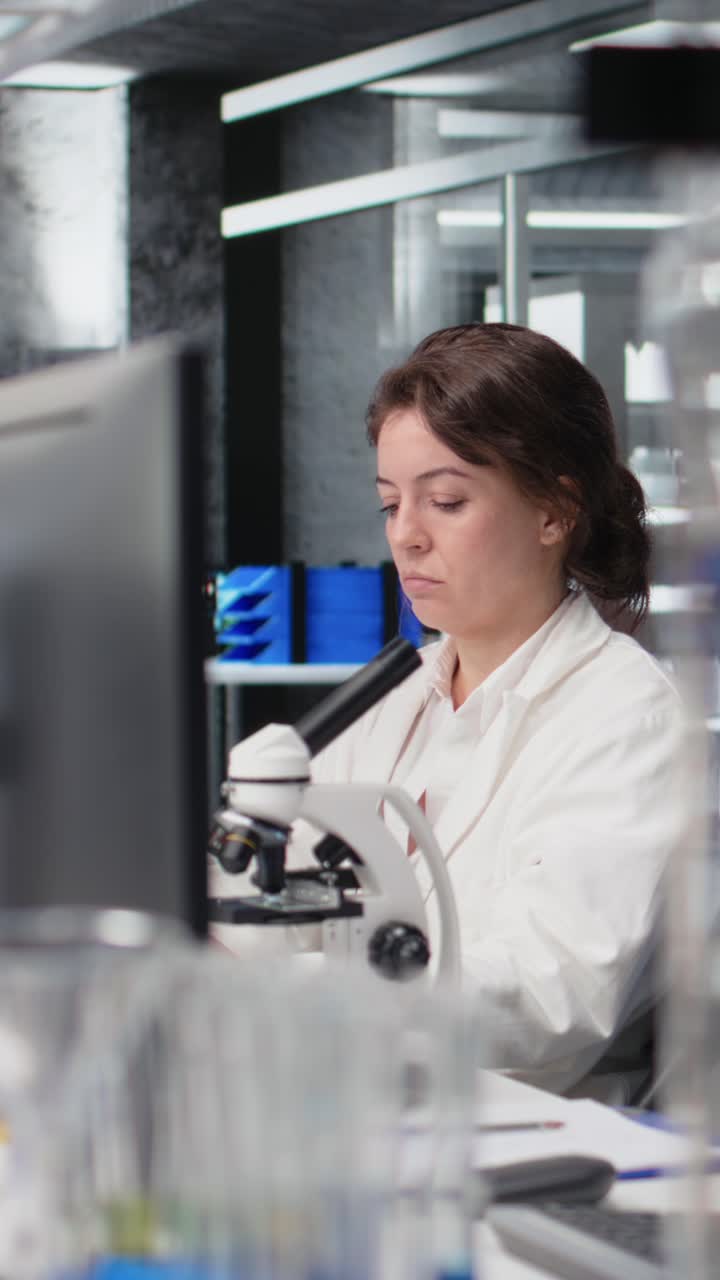 Vídeo vertical de um cientista de laboratório fazendo brainstorming de ideias em uma mesa de computador, enquanto coça o queixo com uma caneta