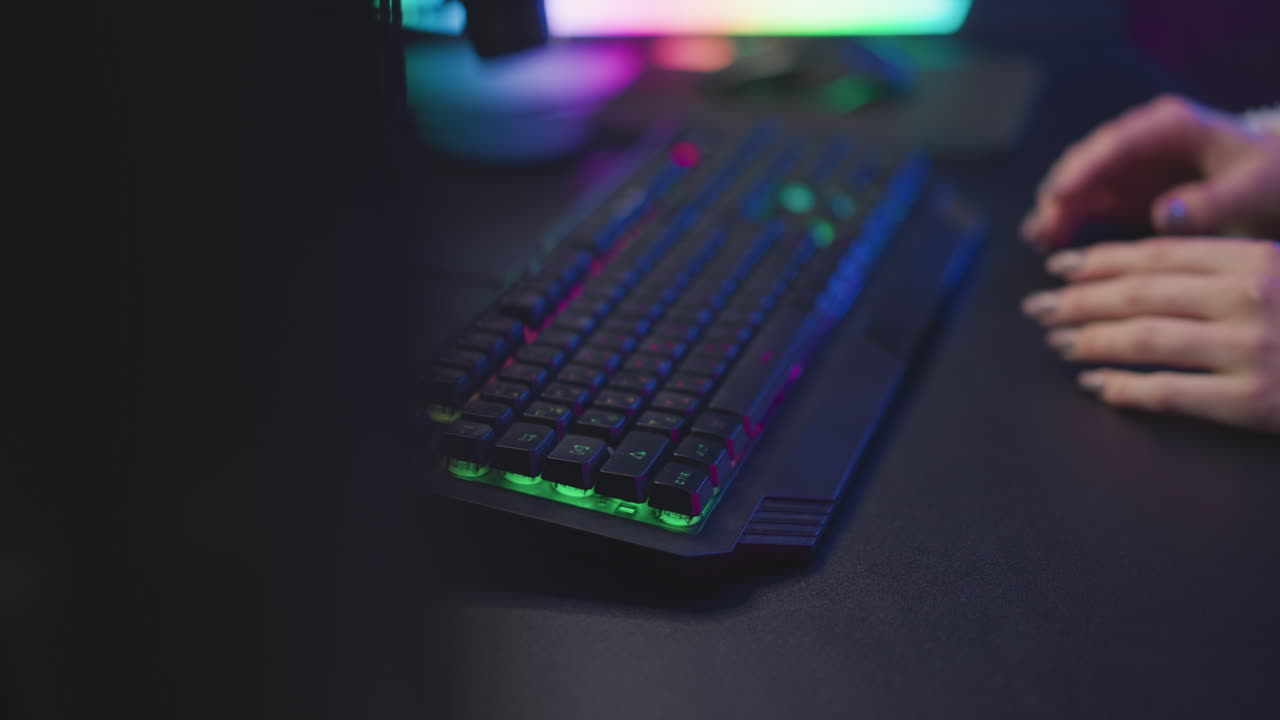 Hand view of lady in fluffy sweater adjusting rgb keyboard close to desktop screen with vibrant color reflections, fingernails painted, fingers poised for typing under colorful ambient lighting