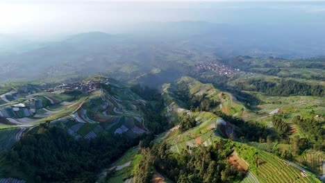 aerial view of terraced farmland on rolling hills with a small rural village nestled on the ridge, surrounded by lush green vegetation under soft morning light. indonesia rural landscape