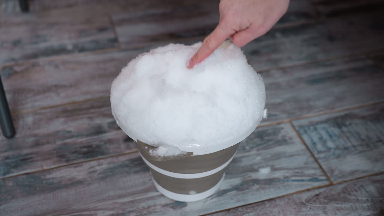 Vessel heaped with fresh snow on tile floor as female fingertips gently press into powdery mound, melting granules glistening under soft indoor light, wooden dress hem and boots blurred in background