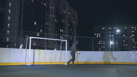 goalkeeper bouncing soccer ball before powerfully kicking it out during practice on an urban field at night, the illuminated backdrop with tall buildings