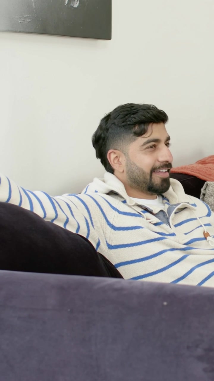 Video verticale: Rispondendo a una voce fuori campo, un uomo con felpa con cappuccio si sistema sul divano di casa con cuscini.