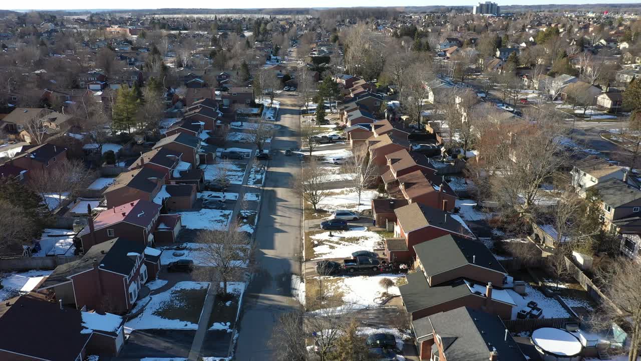 Aerial perspective slowly moving forward above a quiet residential street lined with brick houses and bare trees during a sunny winter day, showing snow covered lawns and a clear cityscape horizon