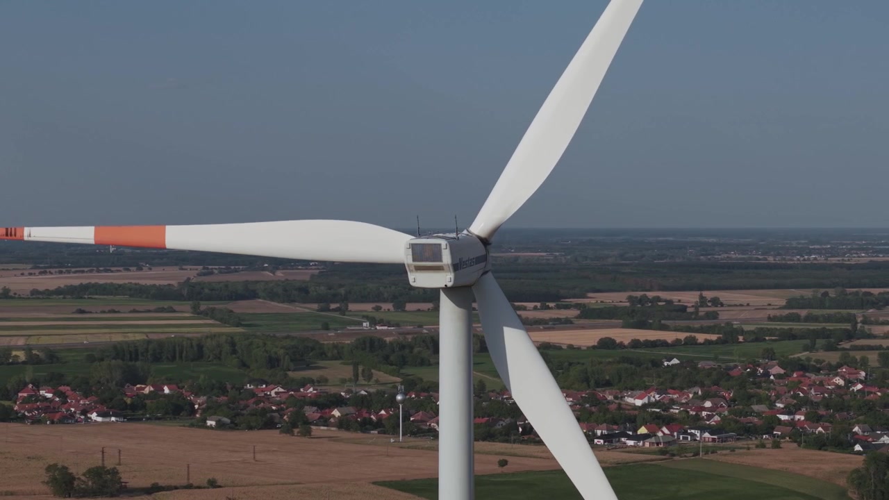 Aerial close-up of a large wind turbine above expansive agricultural fields and a rural village.