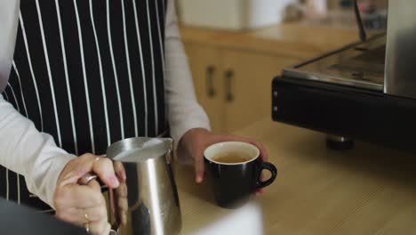 Mittelteil-Einer-Kaukasischen-Kellnerin-Mit-Schürze,-Die-Milch-In-Eine-Tasse-Kaffee-Gießt