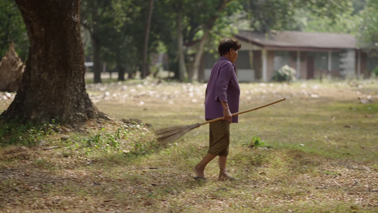 Elderly woman walking with a broom in a rural yard