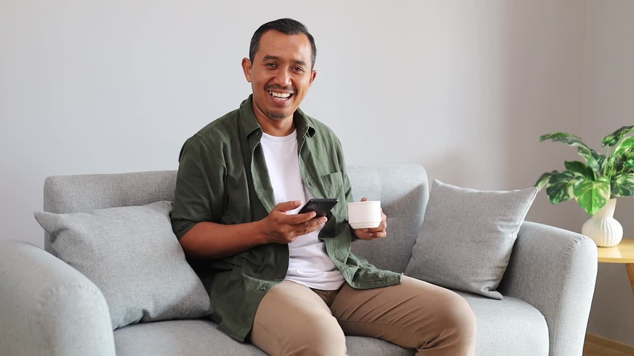 Happy Handsome Asian Man Using Smartphone in Living Room at Home, Resting on Comfortable Sofa. Browsing the Internet and Checking Videos on Social Networks and Having Fun while holding a cup of coffee