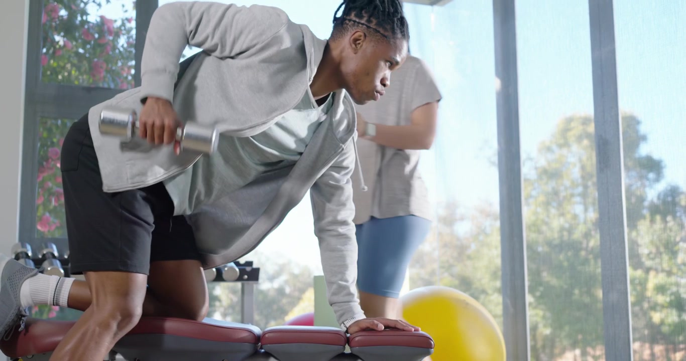 African American man rowing dumbbell at gym bench as Asian coach counting correcting form for power