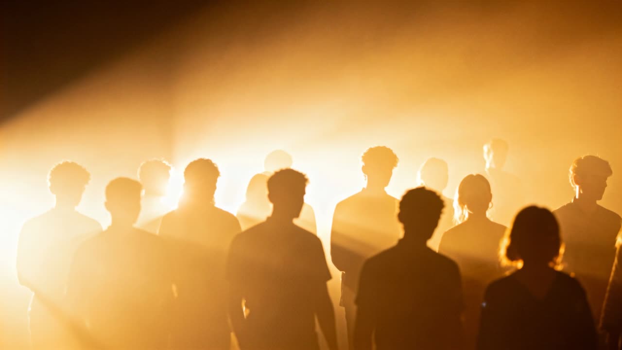 Grupo de pessoas se movendo no palco, reagindo a uma iluminação dourada ampla, com holofotes e névoa.