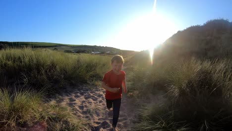 pov shot of young boy on beach vacation running in sand dunes against flaring sun