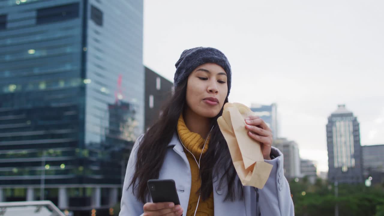 Asian woman walking using smartphone and eating sandwich