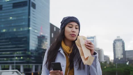 Mujer-Asiática-Caminando-Usando-Un-Teléfono-Inteligente-Y-Comiendo-Sándwich