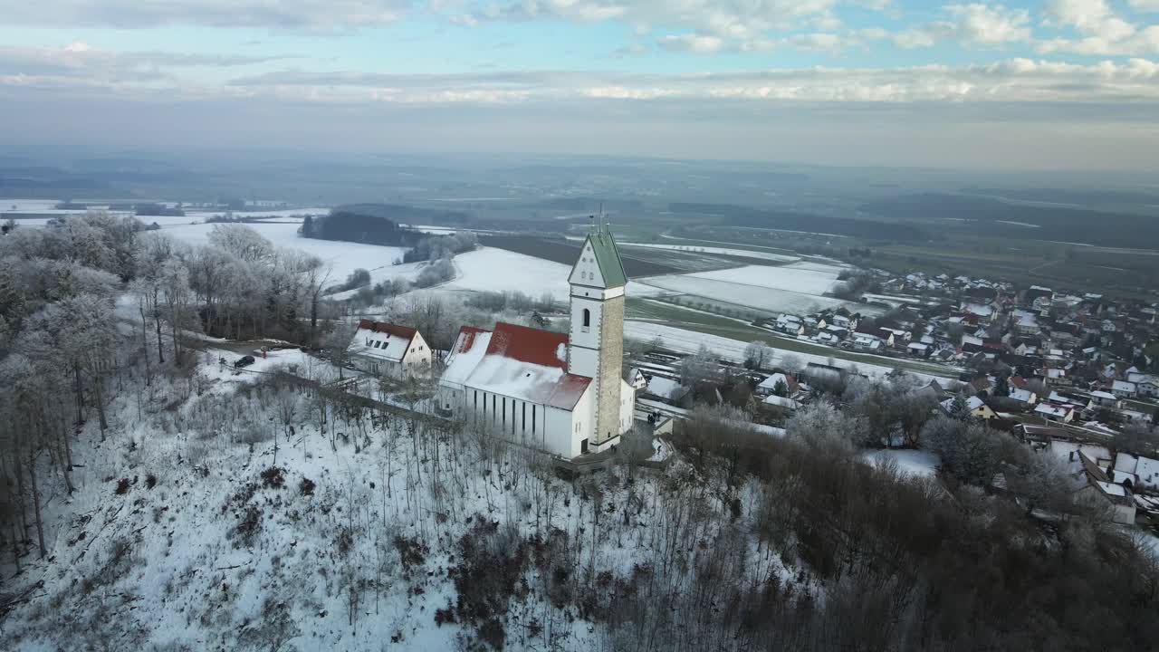 A breathtaking drone shot of a snowy winter landscape featuring a church atop a hill. The drone moves from left to right, revealing snow-covered villages below and a serene backdrop.