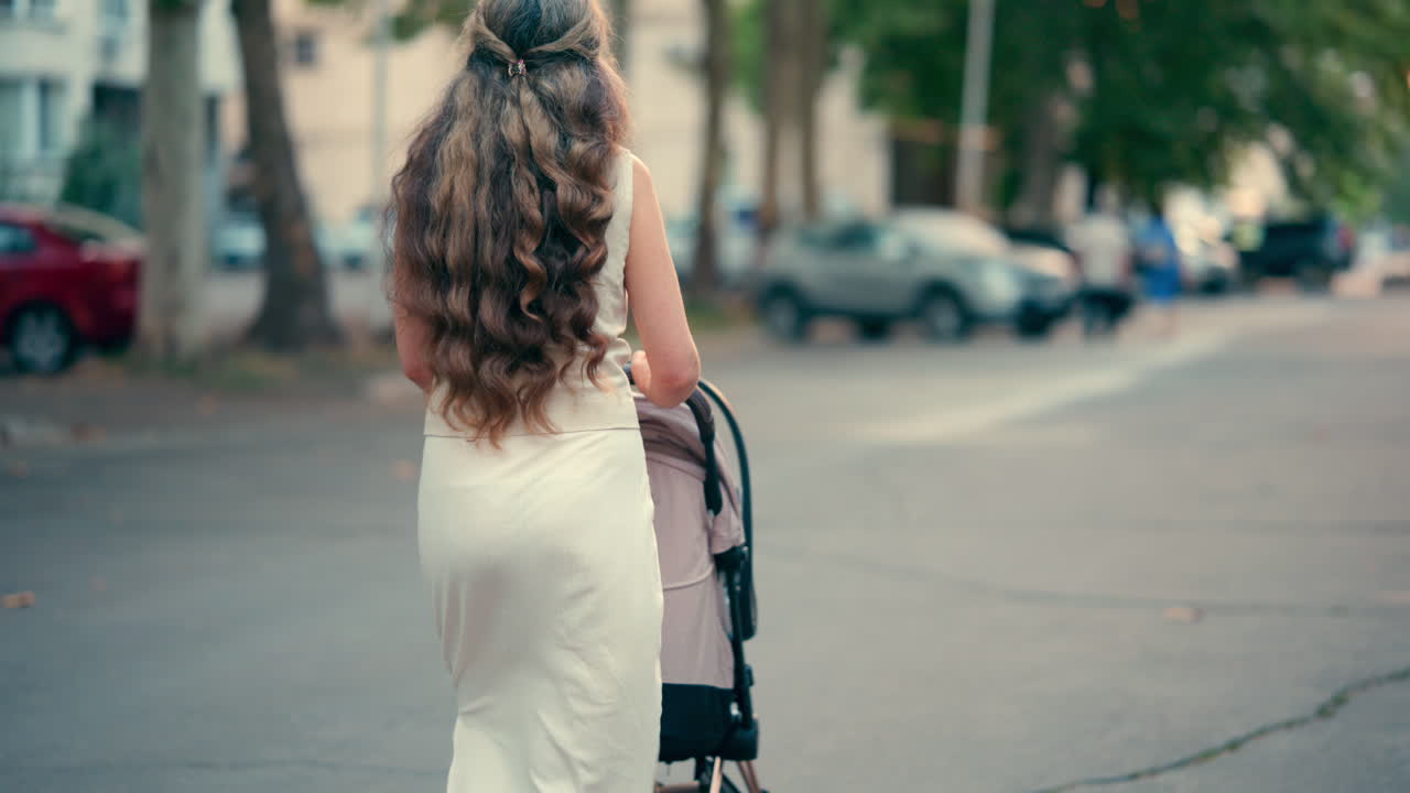 View from the back of a woman with long wavy hair pushing a stroller along a tree-lined park pathway