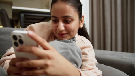 Close-up-a-happy-brunette-girl-with-red-lipstick-in-a-light-jacket-hugs-a-gray-pillow-lying-on-the-sofa-and-types-on-her-smartphone-during-the-day