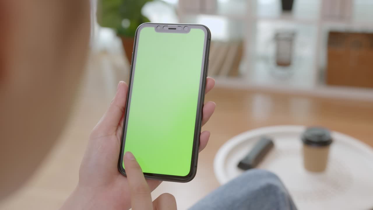 Donna che tiene in mano uno smartphone mockup con schermo verde con messaggio digitato sul divano di casa